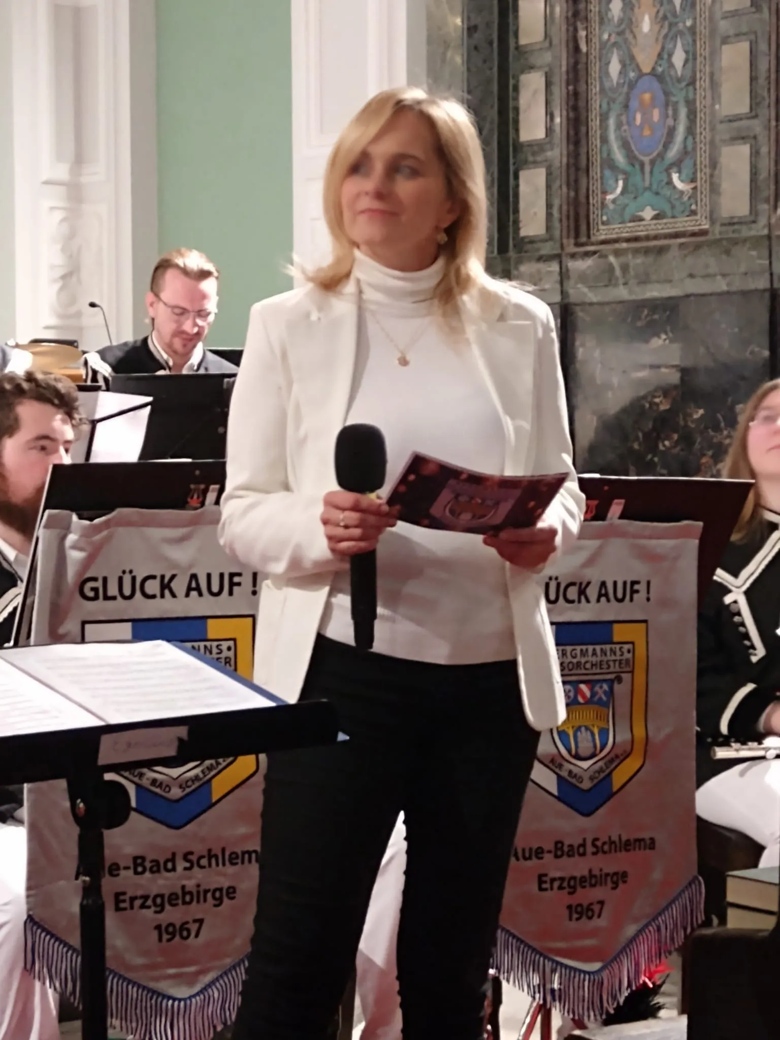 Susanne moderiert im weißen Blazer vor einem Blasorchester